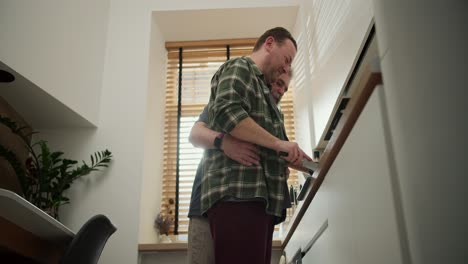 Happy-middle-aged-brunette-man-with-stubble-in-a-green-checkered-shirt-prepares-dinner-while-his-elderly-boyfriend-with-a-gray-beard-and-lush-white-hair-hugs-him-from-behind-in-the-kitchen-during-the-day