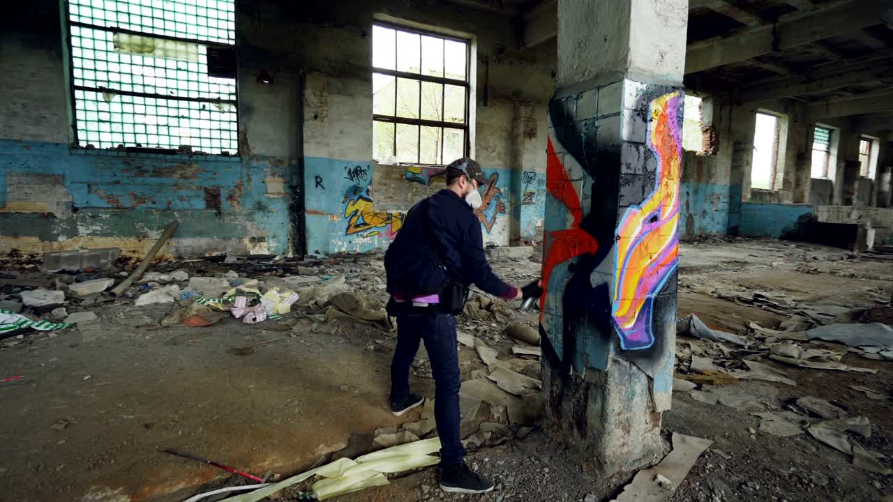 Male graffiti artist is decorating old damaged column inside empty industrial building with abstract pictures. Modern painter is using aerosol spray paint.