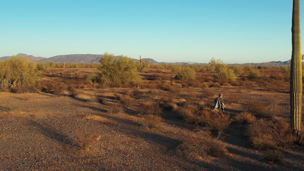 Un desperado del sud-ovest che indossa un poncho cammina attraverso la vegetazione desertica e oltre imponenti cactus Saguaro nel deserto di Sonora al tramonto