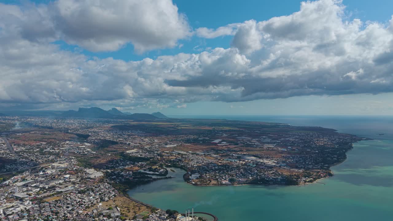 Expansive aerial view of Mauritius island landscape with coastal lagoons, towns, and distant mountains under dramatic tropical clouds and sunlight