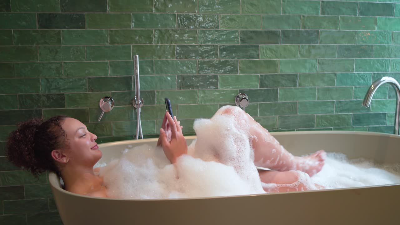 Donna sdraiata in una vasca ovale tra le bolle, con piastrelle verdi e telefono angolato per un selfie, che tiene in mano un barattolo