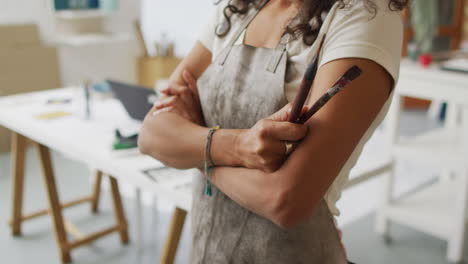 artist in a studio, arms crossed holding a paintbrush