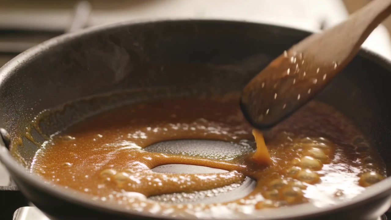 Stirring hand moving wooden spoon bubbling caramel sauce in pan on lit burner, preventing burning