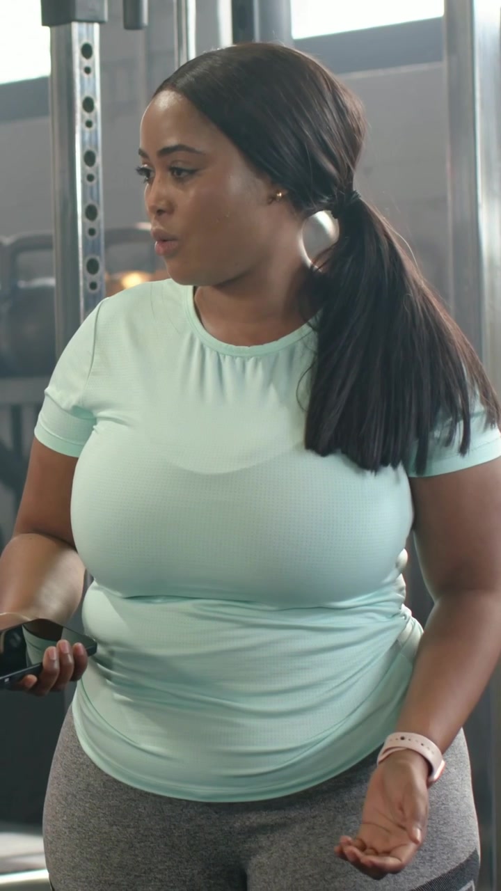 Video verticale: Donna che parla, con un top color menta, controlla il telefono vicino a uno scaffale, rispondendo a una voce prima dell'allenamento.