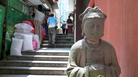 Fußgänger-Gehen-In-Einer-Schmalen-Straße-Die-Treppe-Hinauf-Und-Kommen-An-Einer-Steinernen-Buddha-Statue-In-Einem-Viertel-Vorbei,-Das-Für-Seine-Zahlreichen-Antiquitätengeschäfte-Bekannt-Ist