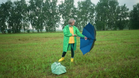 middle-aged woman in green raincoat standing in grassy field closes her umbrella and drops it near her bag on the ground, vast field with trees in the distance and cloudy sky