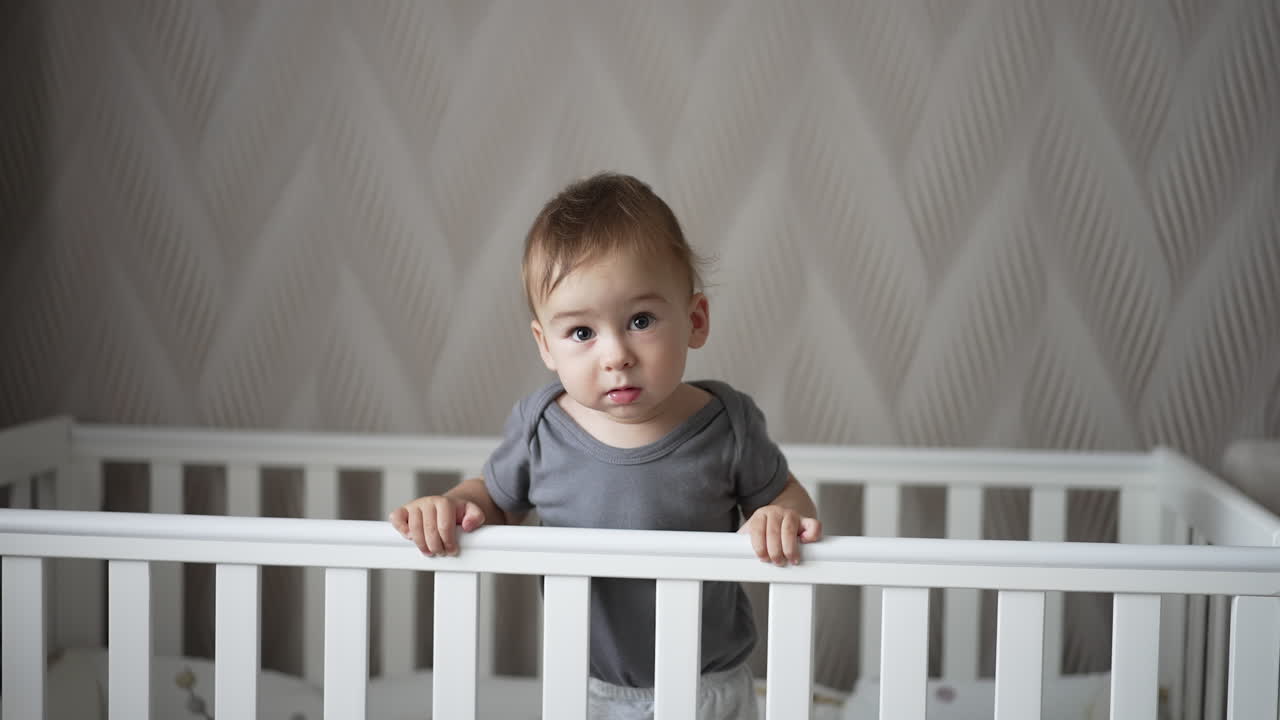 Calm Caucasian baby boy standing in a crib calmly. Child looks into camera and then smiles adorably. Light backdrop.