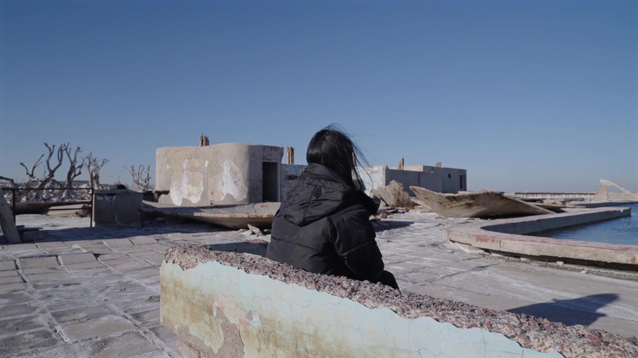 Donna seduta a Villa Epecuén, Buenos Aires, Argentina, rovine di una città allagata con superfici ricoperte da croste di sale, strutture crollate e vegetazione morta che mostrano un ambiente post-disastro, ripresa orbitale con drone.