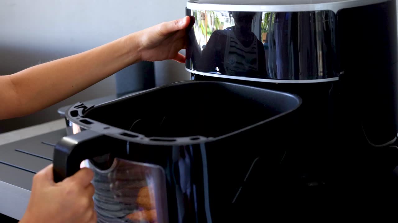 A person operates an air fryer, removing cooked food in a sleek, modern kitchen setting with natural lighting
