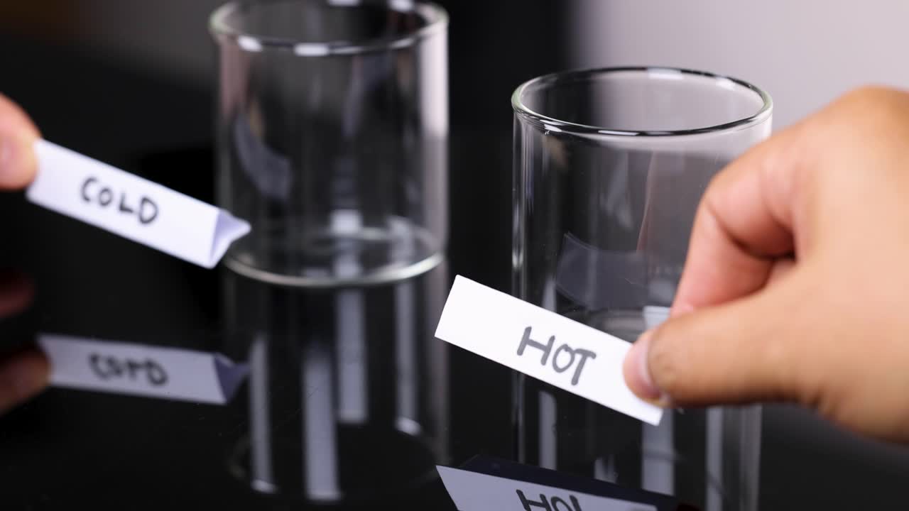 Hand places 'cold' and 'hot' paper labels on two empty glass cups, studio lighting, close-up