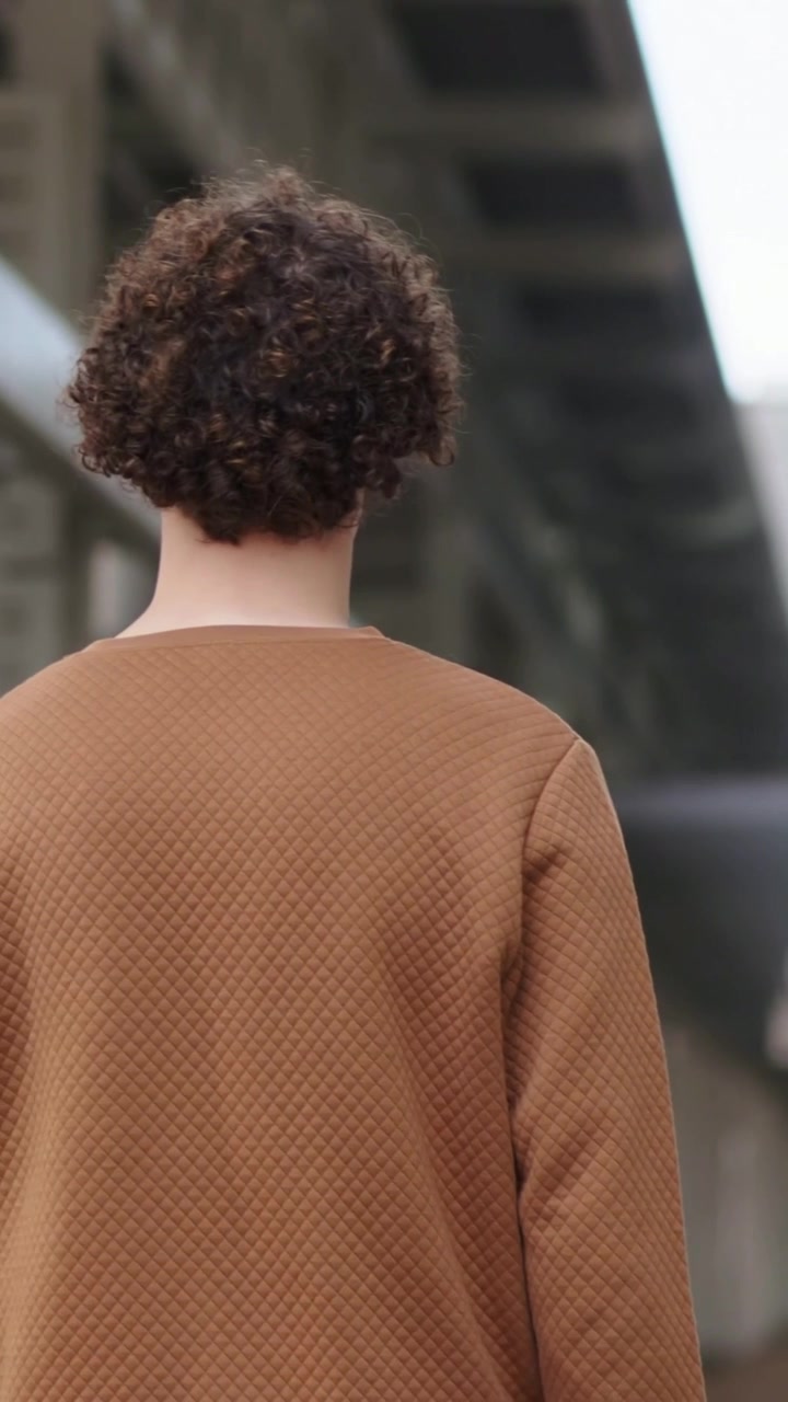 Vertical video: Walking man in brown sweater moving rightward on covered platform, passing skylight