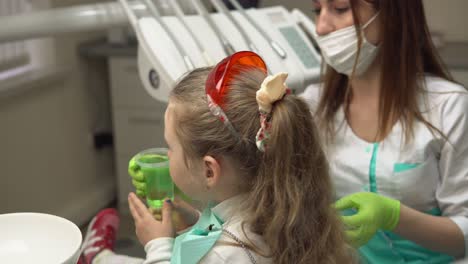 una niña linda enjuagándose la boca con un tratamiento en el dentista. una niña pequeña sosteniendo un vaso con líquido verde en su mano. filmado en 4k