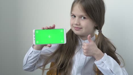 school girl in white blouse shows phone with green screen mock up cellphone, mobile, telephone. chroma key green screen smartphone set up for advertising. education, technology, gadget and kid