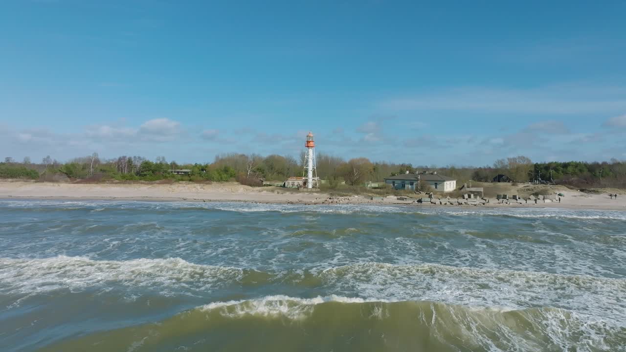 vista aérea del faro de color blanco, costa del mar báltico, letonia, playa de arena blanca, grandes olas, día soleado con nubes, toma de órbita de drones a baja distancia