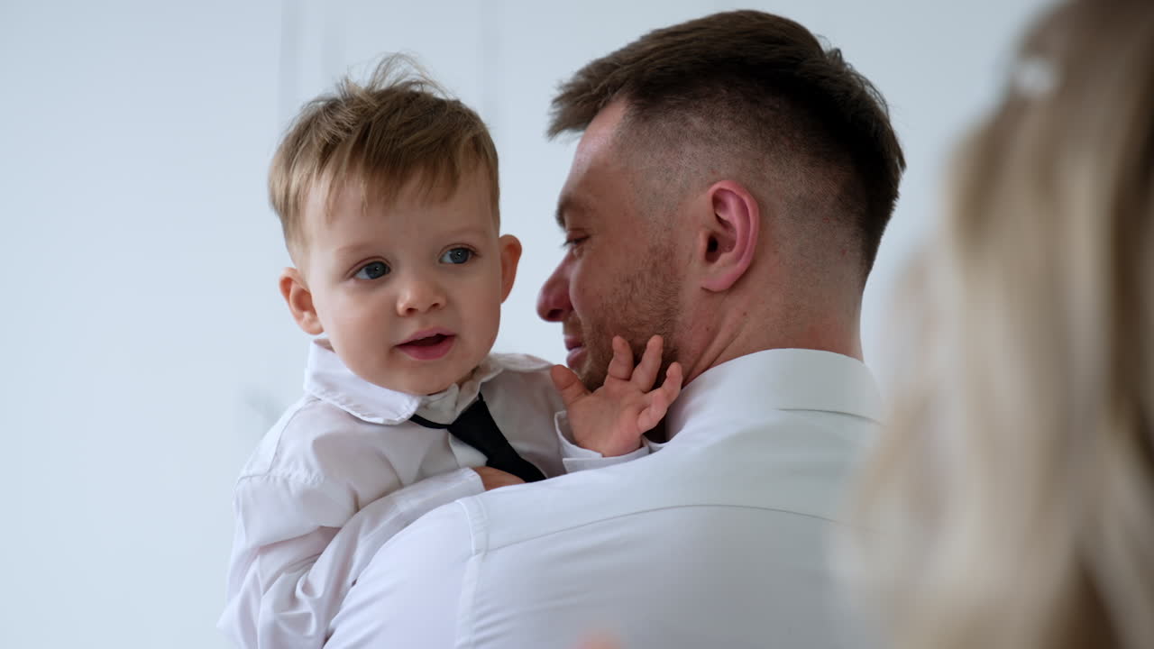 Caucasian man holds a little baby boy in white shirt and black tie. Lovely kid looks at mom standing at foreground.