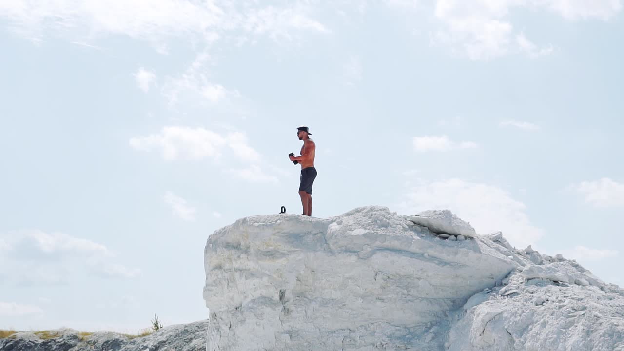 Fitness man drinks water from a bottle on a white mountain on a background of blue sky. Slow motion