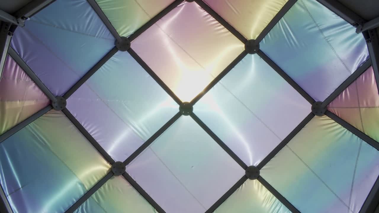 Shifting iridescent panels casting color bands under canopy, moving light revealing metal hubs