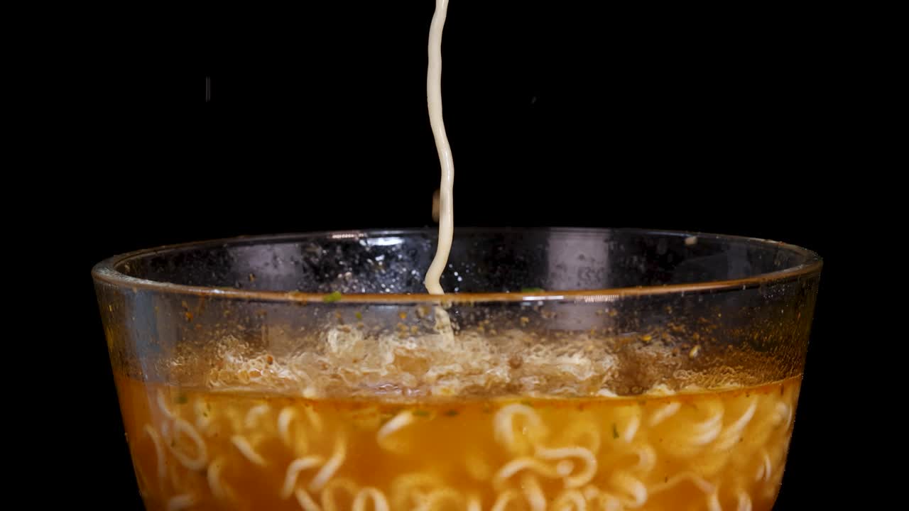 A hand lifts cooked instant noodles from a clear glass bowl filled with broth against a black background, with dramatic studio lighting and a close-up perspective