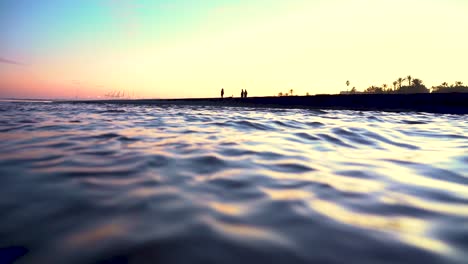 Wasser-Fließt-Mit-Voller-Geschwindigkeit,-Während-Sie-Am-Horizont-Die-Schatten-Von-Menschen-Sehen,-Die-In-Einem-Wunderschönen,-Orangefarbenen-Und-Farbenfrohen-Gegenlicht-Am-Strand-Bleiben