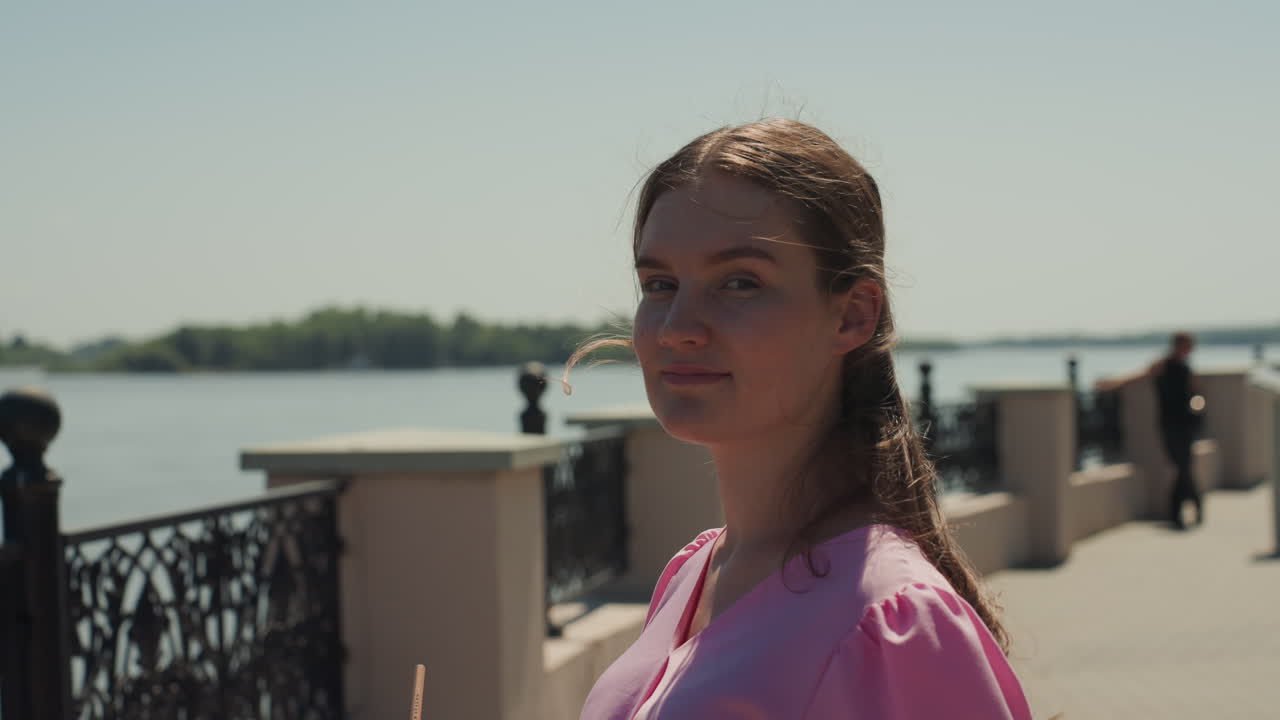 Donna in abito rosa sul lungofiume, tranquilla passerella sul lungomare con ringhiera in ferro battuto, lampioni vintage, tonalità da film delicato, espressione contemplativa, brezza leggera, sguardo spontaneo, estate urbana