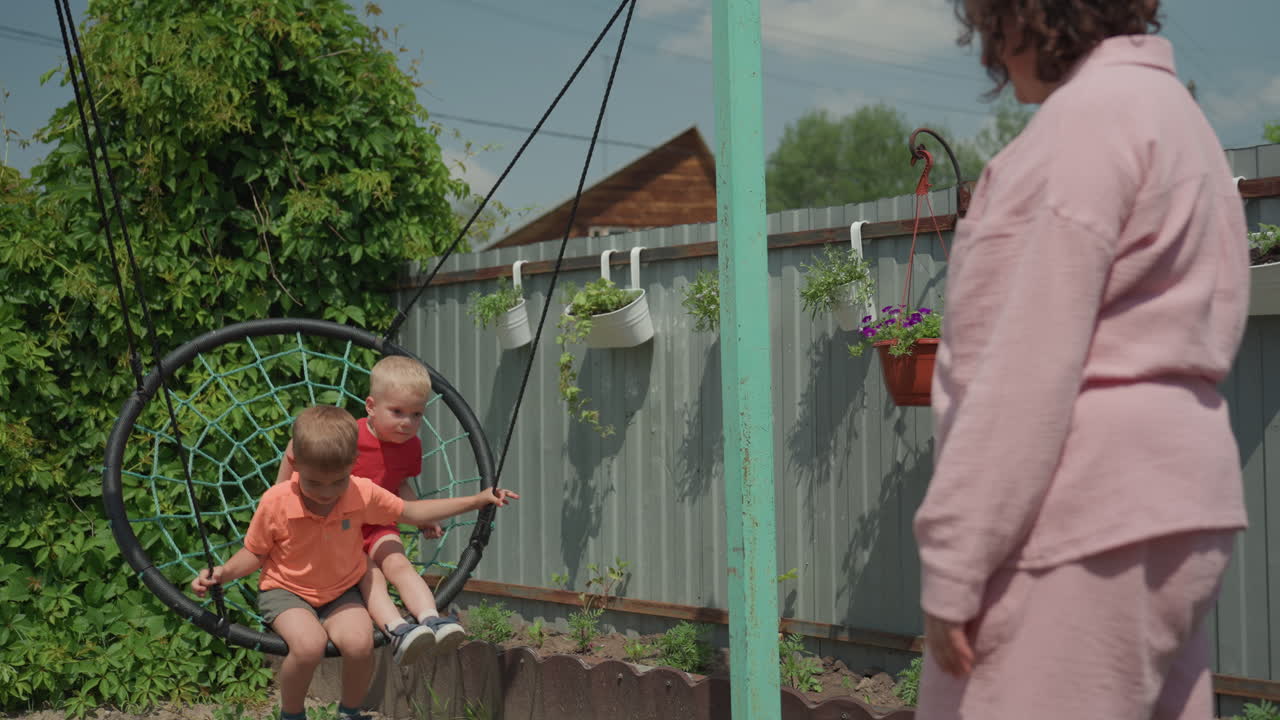 Child On Swing Beneath Bright Summer Leaves, Mother Delights In Pushing Son On Swing During Sunny Day, Happy Mother And Child Enjoy Playful Moments Under Colorful Summer Trees Outdoors
