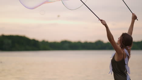 a young hippie woman in a dress and with feathers on her head makes huge soap bubbles at sunset on the shore of a lake in slow motion