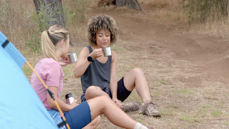 two women are enjoying a camping trip, sitting by a tent with mugs in hand, with copy space