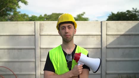 a construction worker, clad in a yellow hard hat with standard safety spectacles hanging from the neckline of his shirt, is gripping a megaphone - medium close up