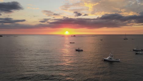 Puesta-De-Sol-Naranja-Brillante-Sobre-El-Horizonte-Del-Océano-Con-Nubes-Y-Reflejo-En-El-Agua-Con-Barcos,-4k-Drone-Costa-Rica-Atardecer