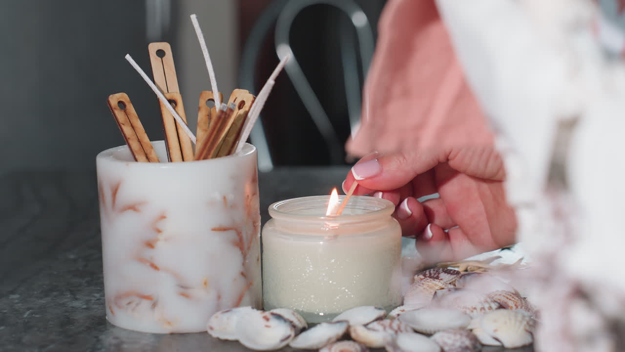 Close up of hand with polished nails lighting white candle with matchstick on dark marble table surrounded by seashells, starfish, wooden sticks in white holder