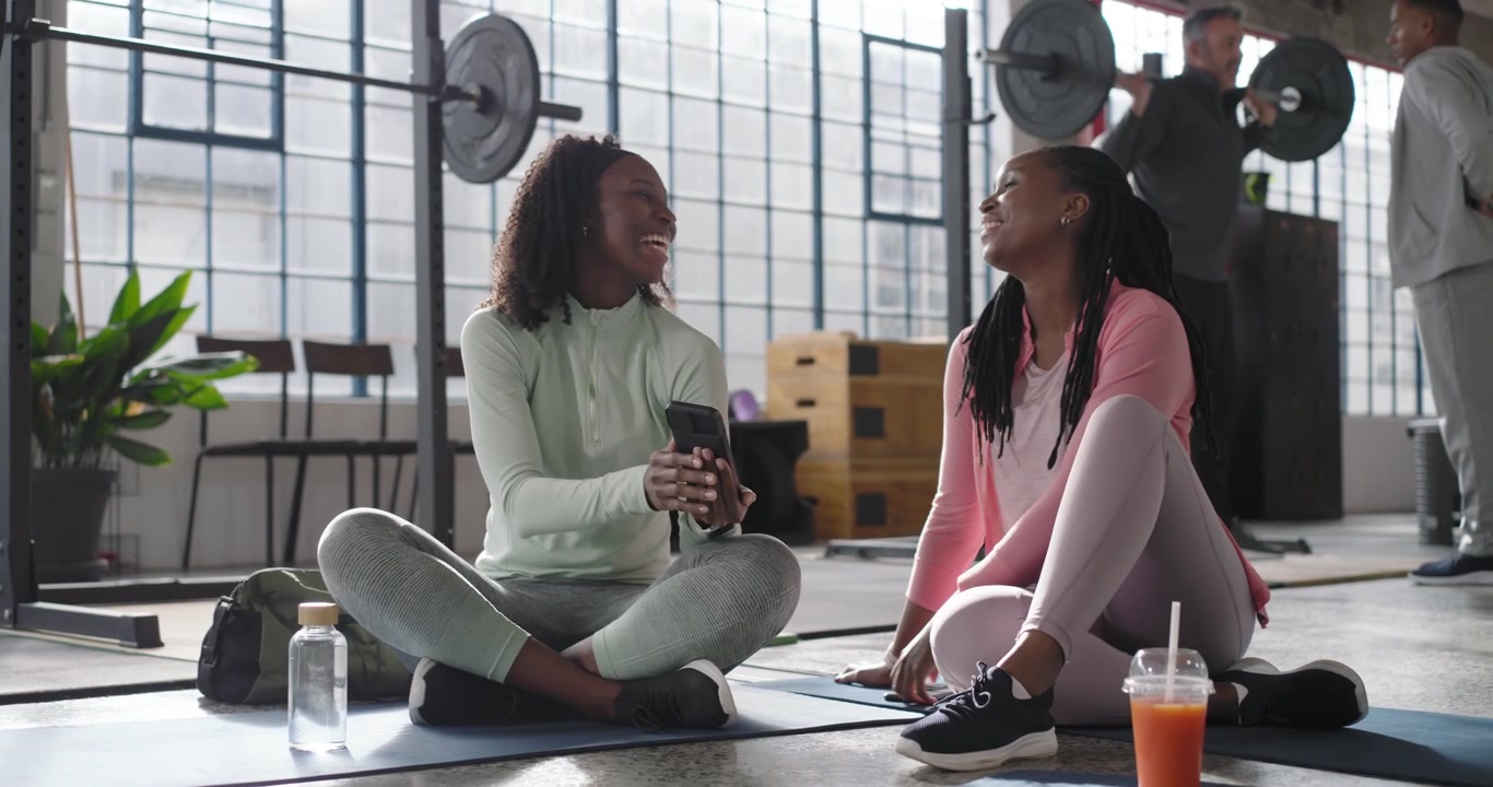 African American female friends sitting on yoga mats in gym, showing phone to share workout clip