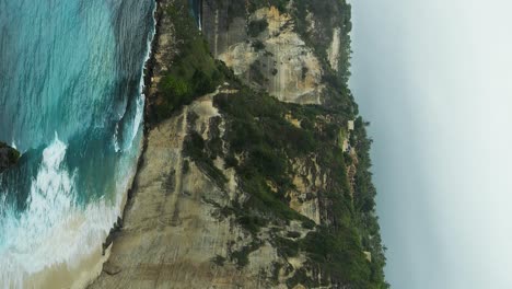 Vertical-slow-motion-panorama-shot-from-the-stunning-Diamond-Beach-on-Nusa-Penida,-Bali,-offering-a-beautiful-view-of-the-imposing-cliffs-and-the-azure-sea,-adorned-by-the-rhythmic-waves