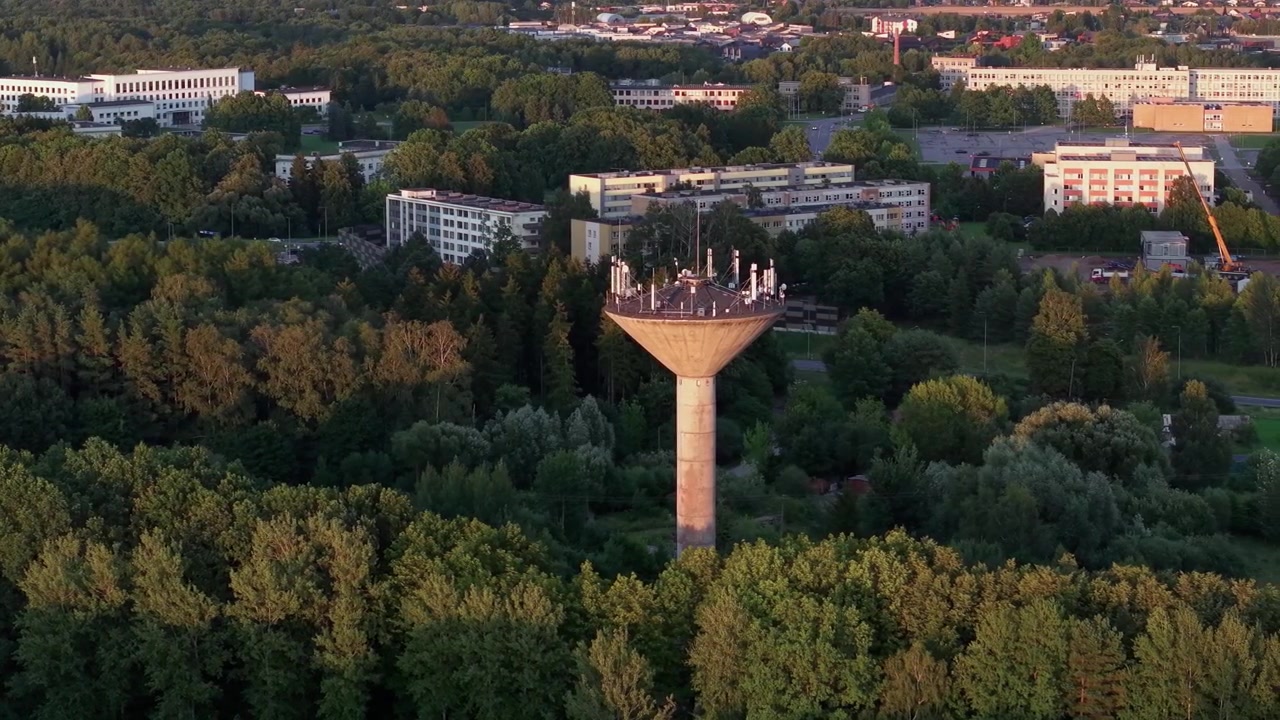 Vídeo de drone de uma alta torre de água de concreto com antenas elevando-se acima de densa floresta verde e edifícios modernos da cidade, capturado sob a luz dourada do pôr do sol e movimento panorâmico aéreo