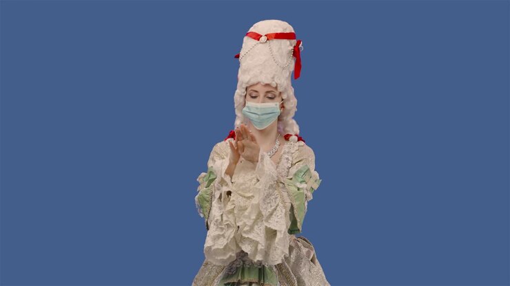 Portrait of courtier lady in vintage dress, wig and medical mask treats his hands with an antiseptic. Young woman posing in studio with blue screen background. Close up. Slow motion ready 59.94fps