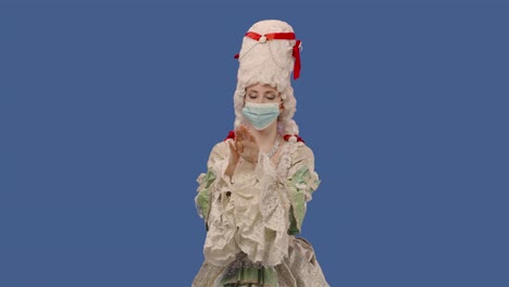 portrait of courtier lady in vintage dress, wig and medical mask treats his hands with an antiseptic. young woman posing in studio with blue screen background. close up. slow motion ready 59.94fps