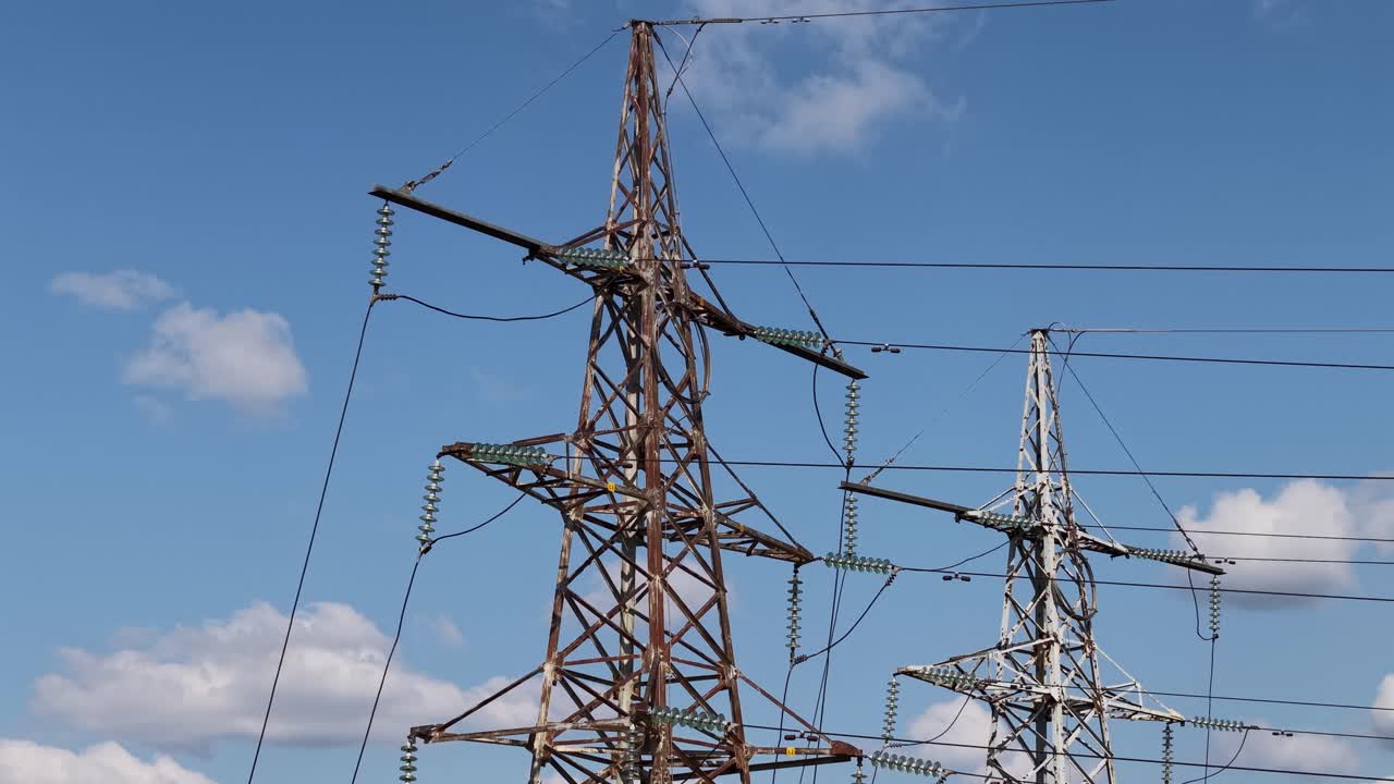 Two high-voltage transmission towers and power lines against a bright blue sky with scattered clouds, showing energy grid infrastructure in clear daylight