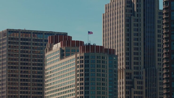Arranha-céus do centro de Chicago subindo em um céu azul aberto com uma bandeira nacional hasteada — uma metáfora visual para poder corporativo, ambição e força econômica