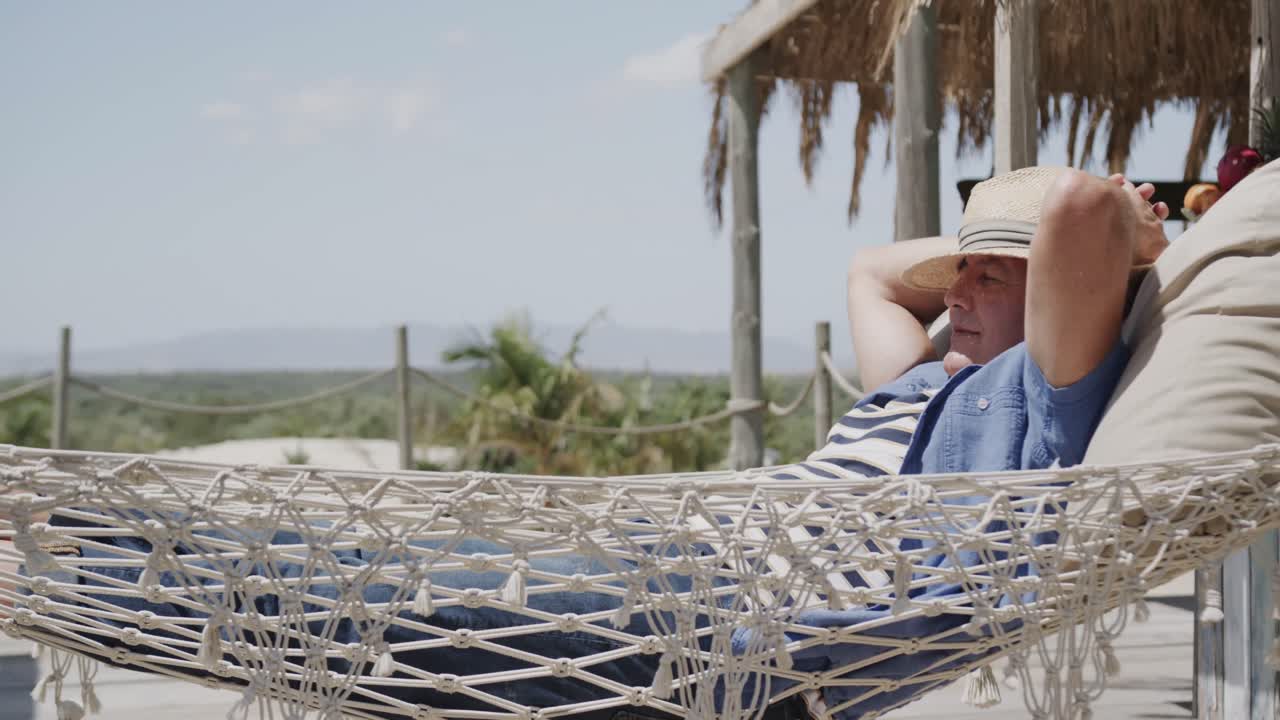 hombre caucásico mayor feliz con sombrero relajándose en una hamaca al sol en el bar de la playa, en cámara lenta