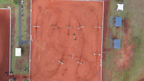 aerial view of a red dirt equestrian jumping arena showing fences, course layout, and hoof track patterns from above, wide symmetrical shot, used for training or competition, drone top-down
