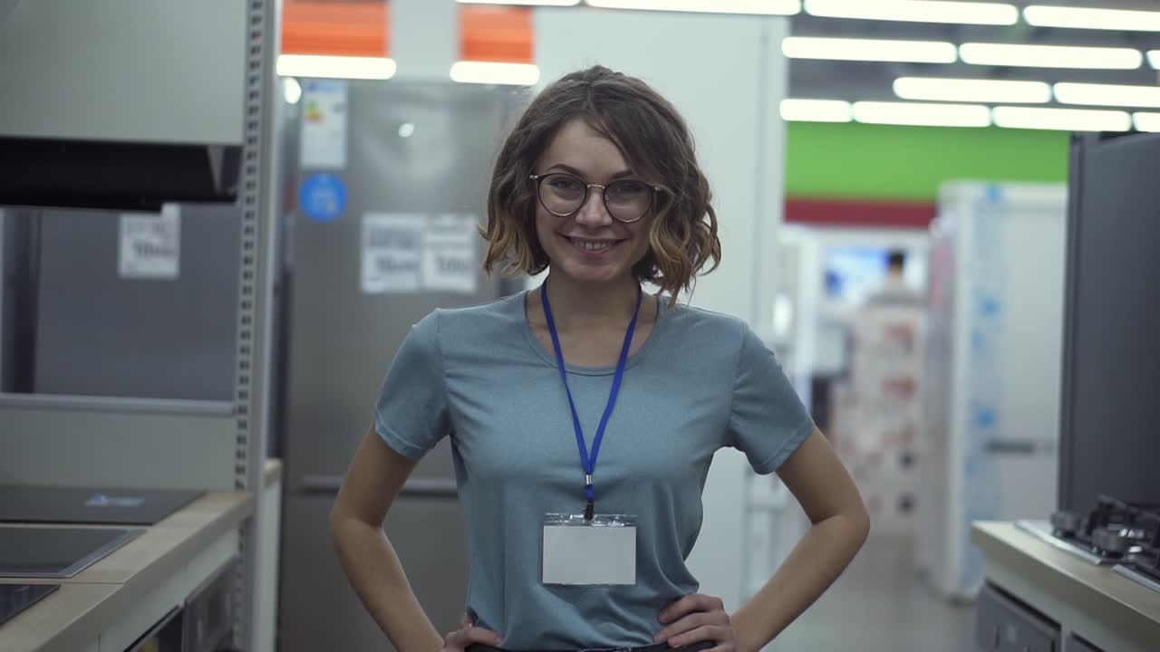 Positive female seller or shop assistant portrait in supermarket store. Woman in blue shirt and empty badge looking at the camera and smiling. Household appliances on the background. Close up camera