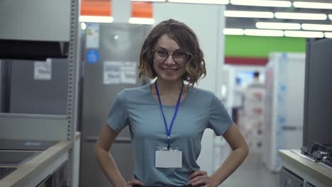 Positive-female-seller-or-shop-assistant-portrait-in-supermarket-store.-Woman-in-blue-shirt-and-empty-badge-looking-at-the-camera-and-smiling.-Household-appliances-on-the-background.-Close-up-camera