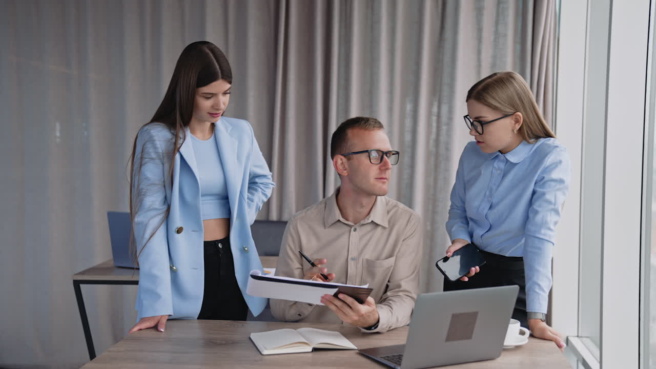 Office people at work in the light room. Man sitting at desk looking through the documents and commenting. His female colleagues standing on both sides of him.