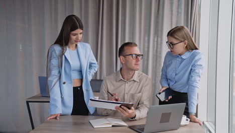 office people at work in the light room. man sitting at desk looking through the documents and commenting. his female colleagues standing on both sides of him.