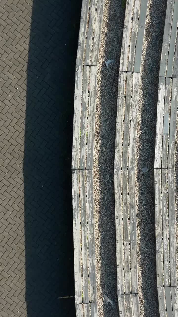 Weathered wooden benches spanning gravel terrain at Sauleskalns amphitheater, revealing textural landscape details of historic Latvian outdoor venue in Talsi, vertical top down drone shot