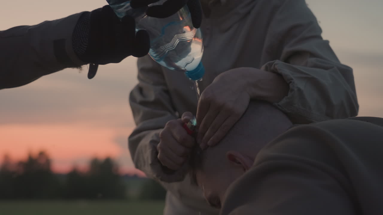 woman pouring water from plastic bottle onto small flame burning on passenger hair while holding lighter close to scalp under pastel sunset sky above grassy field