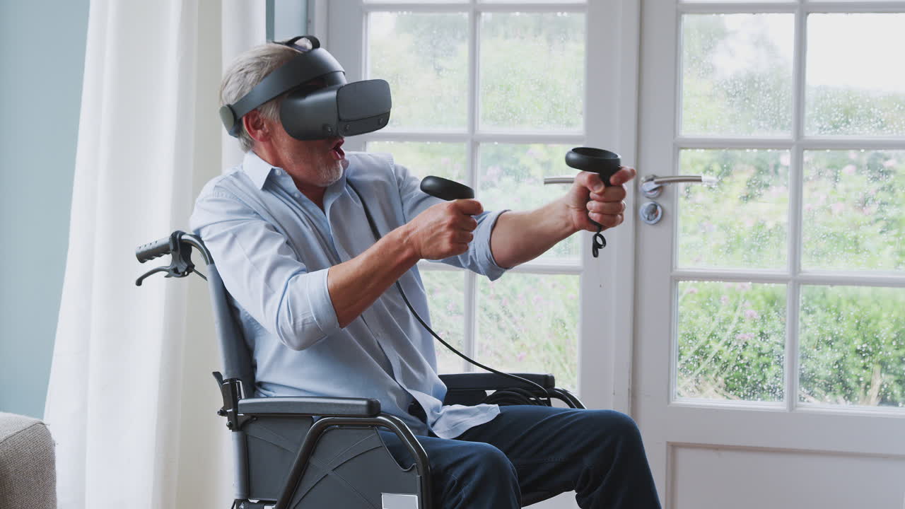 Senior Disabled Man In Wheelchair At Home Wearing Virtual Reality Headset Holding Gaming Controllers