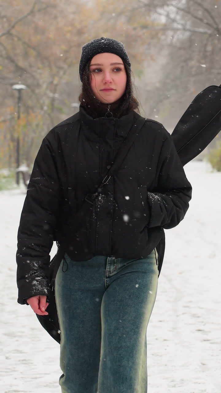 dama con chaqueta de invierno caminando durante la nieve, otra persona siguiendo, camino de parque nevado alineado con postes de lámparas y bancos, escena de invierno tranquila con copos de nieve que caen, árboles que conservan hojas de otoño