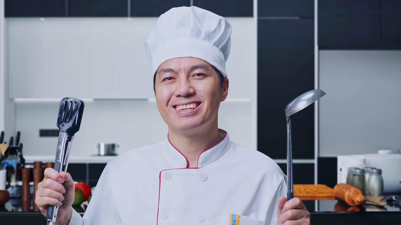 close up de un chef asiático sonriendo y mostrando pinzas y cuchara a la cámara mientras está de pie en la cocina de casa