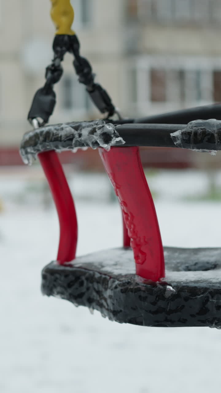 Frozen children's swing coated with ice and icicles hanging from its red and black frame, suspended by yellow ropes, gently swaying against a snowy playground and blurred building background