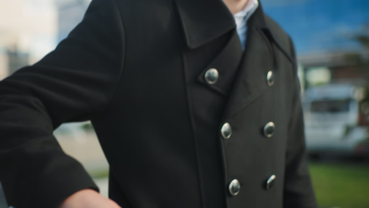 흑색 코트를 입은 남자가 도시 환경에서 야외에서 전화를 취하기 위해 주머니에 손을 고, 들어오는 전화에 응답할 준비가 된 클로즈업, 배경에는 흐릿한 현대 건물과 초록색이 포함되어 있습니다.